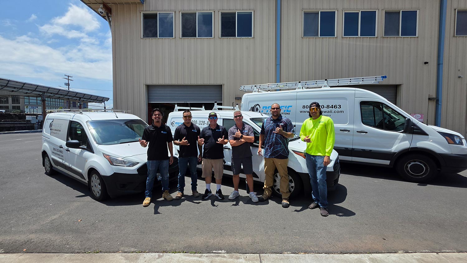 Group of six men smiling and posing next to three white commercial vans outside a building