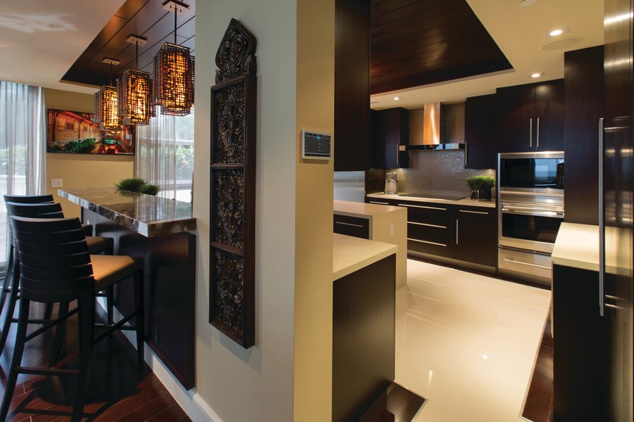 View of a kitchen with dark wood cabinets, hanging lights, and a Control4 panel on the wall. 