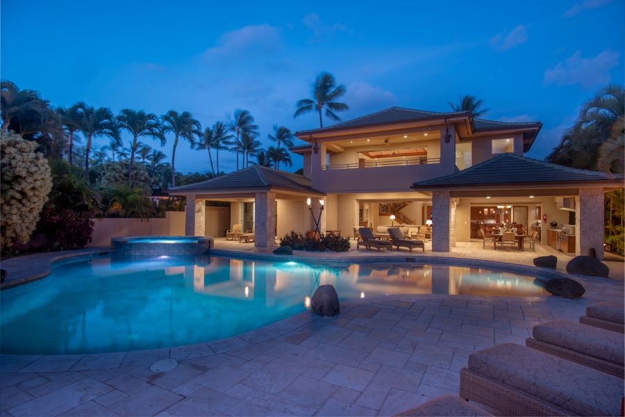 View of the back of a well-lit open-floor plan home across their pool at dusk.