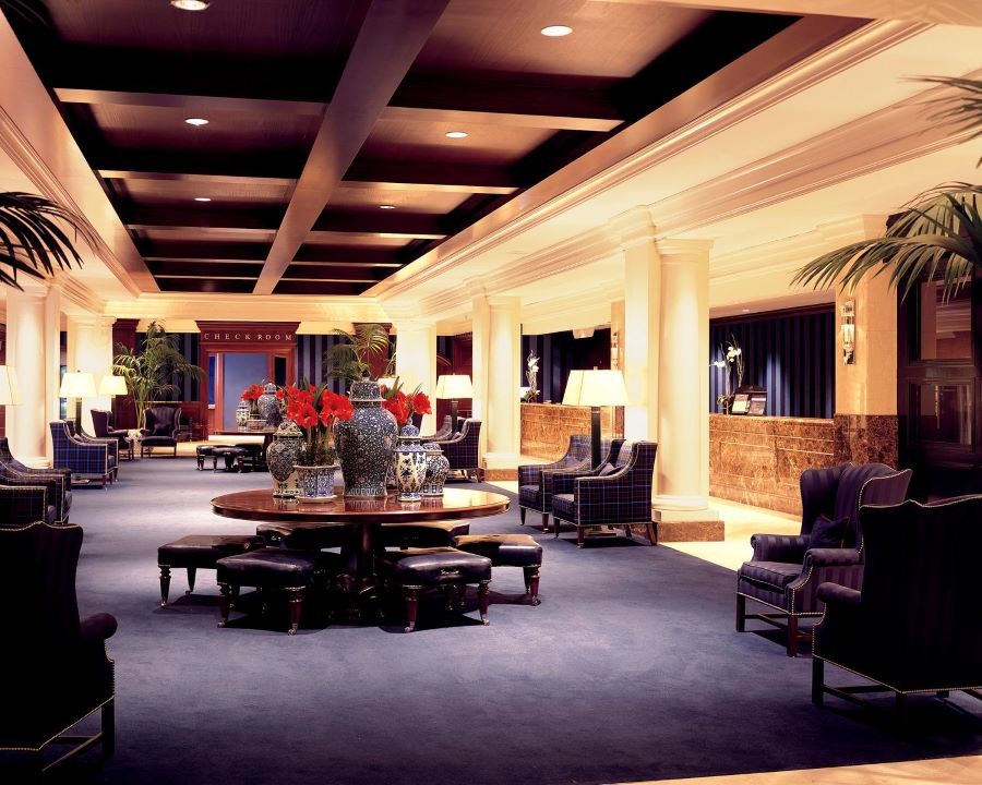 A luxury hotel lobby with plush chairs, vases, a wood ceiling with recessed lights, and the words “Check Room” over a doorway in the background. 