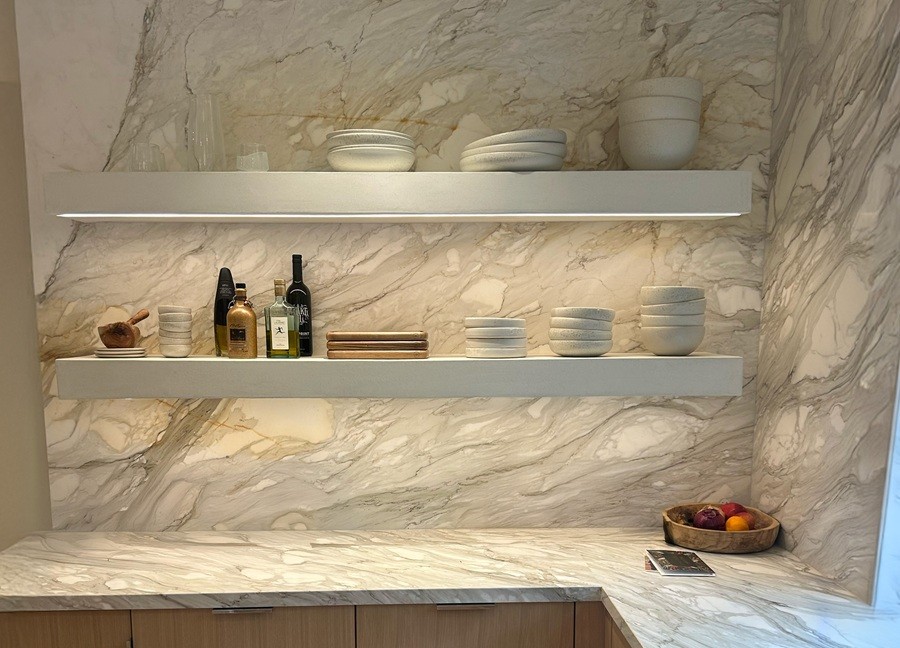 Elegant open kitchen shelves display ceramic bowls, glassware, and bottles against a marble backsplash with under-shelf lighting.