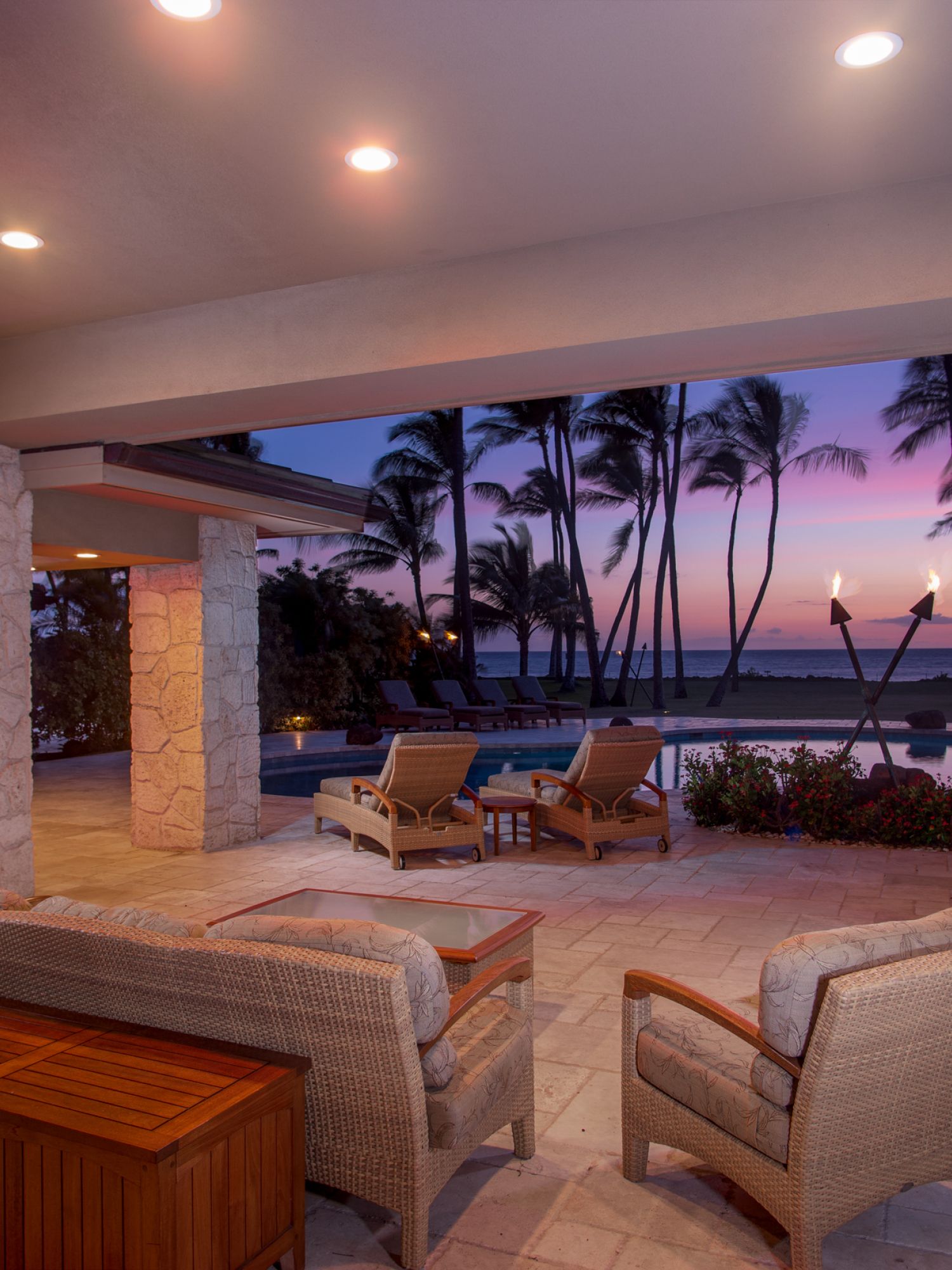 Covered patio with outdoor seating, tiki torches, and pool view at sunset with palm trees